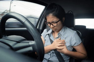 Woman clutching her chest in pain behind the wheel of a car